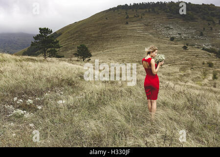 Woman in red dress holding a field flowers while standing in a meadow against terraced slope on a cloudy windy day Stock Photo