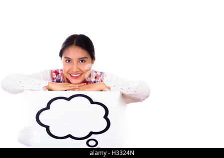 Charming young woman wearing traditional andean blouse with colorful embroideries, matching necklace and earrings, facing camera while posing, leaning over sign which has conversation bubble drawn on it, white studio background Stock Photo