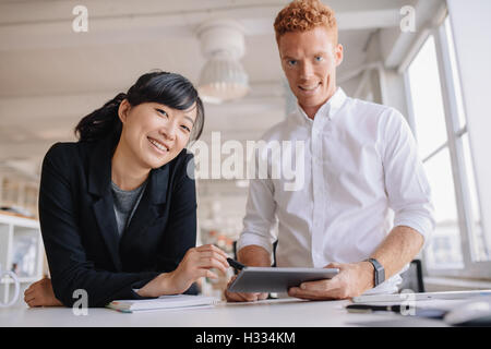 Portrait of successful young business partners standing at a table in office with digital tablet in hand. Businesspeople using d Stock Photo