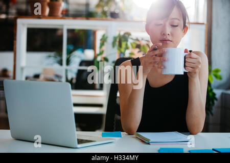 Portrait of relaxed young woman sitting at her desk and having coffee. Asian business woman taking coffee break in office. Stock Photo