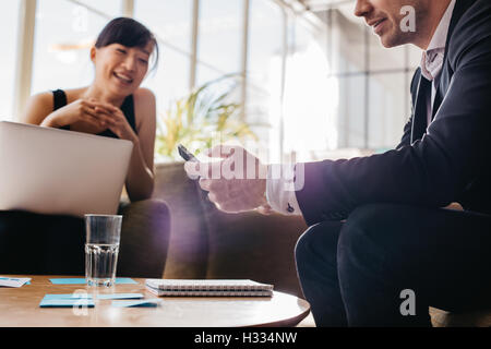 Young business man using mobile phone with woman sitting in background. Business people meeting in office lobby. Stock Photo