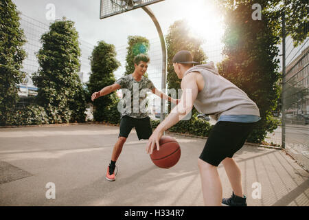 Young friends playing basketball together, boy in front of net blocking and other dribbling the ball on outdoor court.