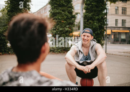 Smiling young guy sitting on basketball court and talking with friend. Two friends relaxing after playing basketball on court.