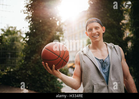 Portrait of smiling young man holding a basketball on outdoor court. Teenage guy looking at camera with a ball in hand.