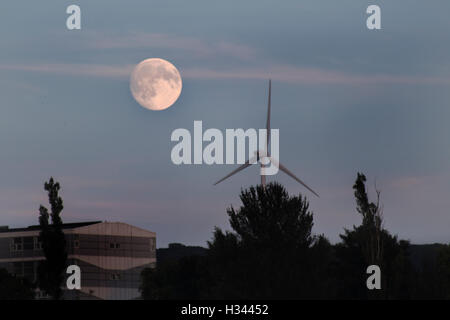 full moon rising over Glasgow, over a building with a wind mill and trees, and blue sky Stock Photo