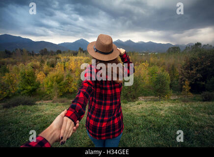 Woman in red checked shirt and hat holding man by hand going to Autumn forest with mountains and cloudy sky Stock Photo