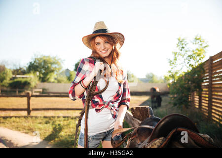 Happy redhead young woman cowgirl preparing saddle for riding horse Stock Photo