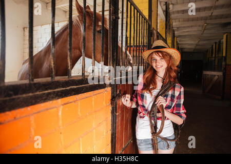 Cheerful attractive young woman cowgirl with horse standing in stable Stock Photo