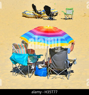 Mature couple under colourful UK beach umbrella relaxing in fold up chairs on sandy British seaside holiday beach shade from hot summer sunshine Stock Photo