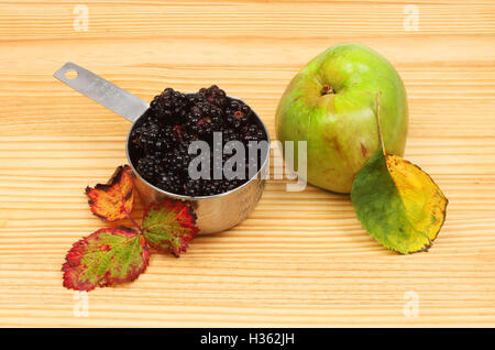 Blackberries in a measuring cup with a cooking apple and Autumnal leaves on a wooden board Stock Photo
