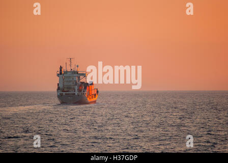 Small container ship sailing across the Agean Sea at Sunset, sailing towards the sun. Stock Photo