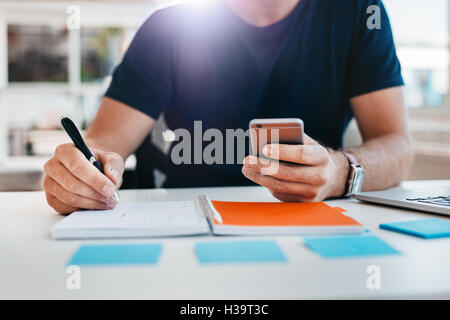 Close up shot of a man taking down note in a personal organizer with mobile phone in other hand. Young man hands working at his Stock Photo