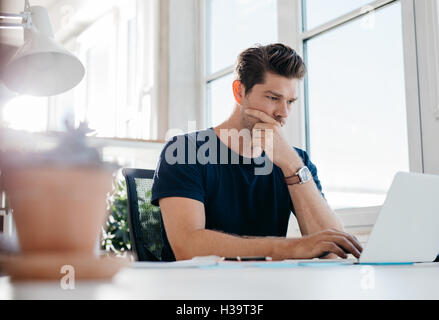 Young man working on laptop in office and thinking. Young male executive using laptop at his desk. Stock Photo