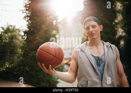 Portrait of handsome young man holding a basketball on outdoor court. Teenage streetball player looking at camera with a ball in Stock Photo