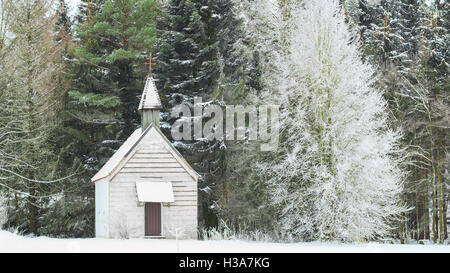 Peaceful winter view of little snow-capped rural wooden church on snowfield glade in frozen forest Stock Photo