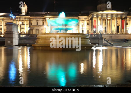 Trafalgar Square and the National Gallery in London at night, with lights and reflections on the fountain surface.