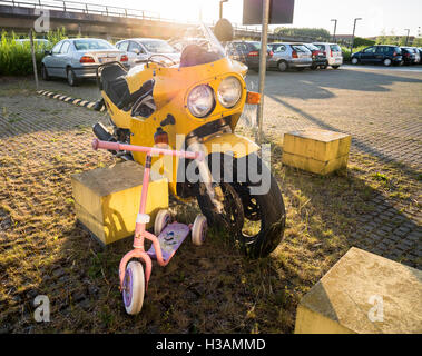 Yellow motorcycle left standing abandoned in a parking lot together with a small kids scooter Stock Photo