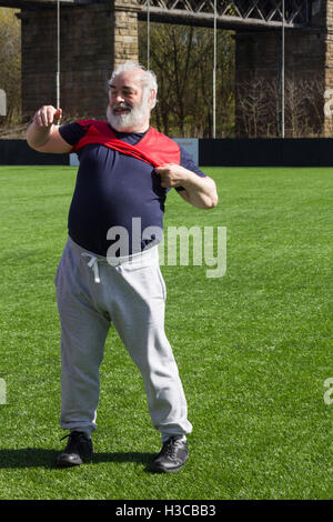 Overweight middle-aged man struggling  to put on a bib shirt ready to take part in a walking  football session. Stock Photo