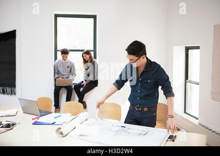 businessman analyzing blueprints while colleagues using laptop in background Stock Photo