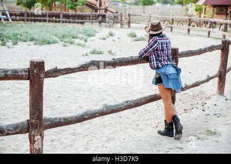 Back view of young woman cowgirl in hat standing near fence on ranch Stock Photo