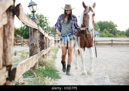 Attractive young woman cowgirl in hat smiling and walking with her horse in village Stock Photo