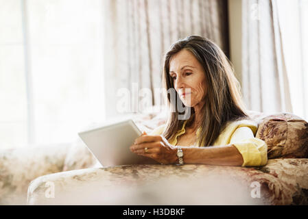 Senior woman with long brown hair sitting on a sofa, using a digital tablet. Stock Photo