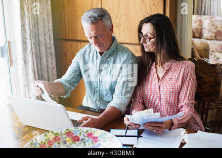 Senior couple sitting at a dining table, using a laptop computer, paperwork and bills on the table. Stock Photo