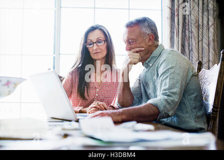 Senior couple sitting at a dining table, using a laptop computer, paperwork and bills on the table. Stock Photo