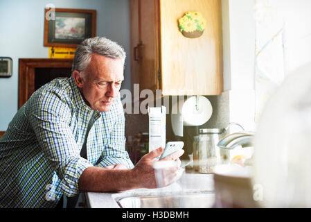 Senior man standing in a kitchen, using a mobile phone. Stock Photo