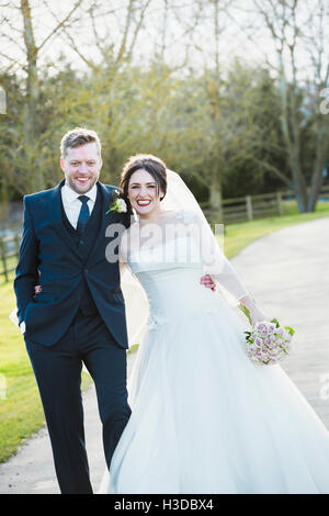A bride and groom on their wedding day walking arm in arm down a path in the sunshine laughing. Stock Photo