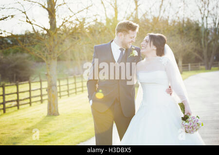 A bride and groom on their wedding day walking arm in arm down a path in the sunshine laughing. Stock Photo