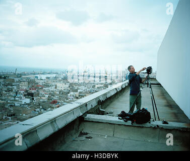 Photographer standing on a rooftop in a city, setting up his camera on a tripod. Stock Photo