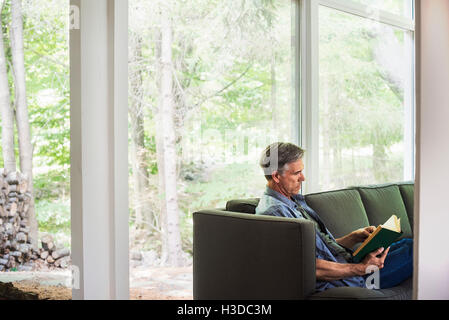 A man sitting by a window on a sofa, reading a book. Stock Photo