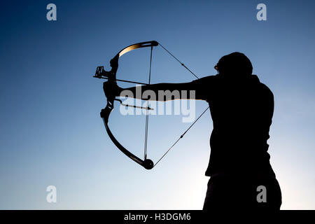 An archer drawing his compound bow in a field in the forest during the early autumn. A silhouette against the clear blue sky in  Stock Photo