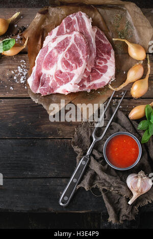 Raw steak meat pork neck on baked paper with shallot onion, tomato sauce marinade, basil and seasoning, served with meat fork ov Stock Photo
