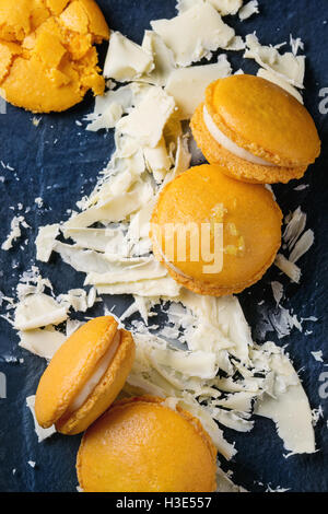 Whole and broken orange lemon homemade macaroons with chopped white chocolate and citrus sugar and zest on dark glass board over Stock Photo