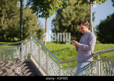 Young hispanic man, blue shirt and jeans, standing on stairs outdoors, holding mobile phone in his hand, smiling - communication concept Stock Photo