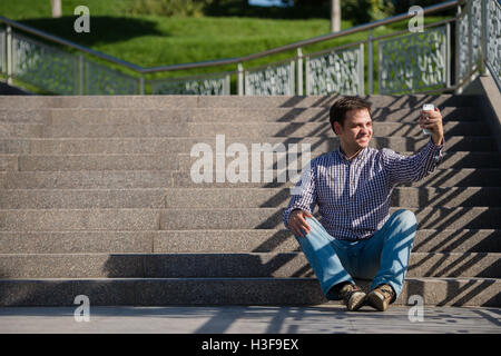 Young, happy man sitting on the stairs, holding mobile phone and making a selfie. Stock Photo