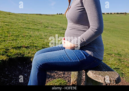 Young pregnant woman in her 20s takes a rest on park bench as she holds her stomach Stock Photo