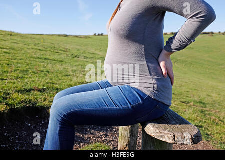 Young pregnant woman in her 20s takes a rest on park bench as she holds her back which aches Stock Photo