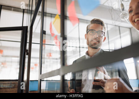 Portrait of young businessman with female colleague looking at adhesive notes on glass wall and brainstorming. Stock Photo