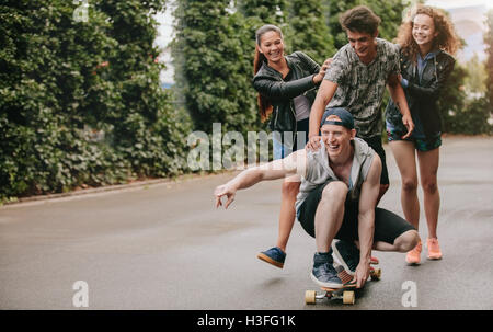 Full length shot of teenage guys on skateboard with girls pushing. Multi ethnic group of friends having fun outdoors with skateb Stock Photo