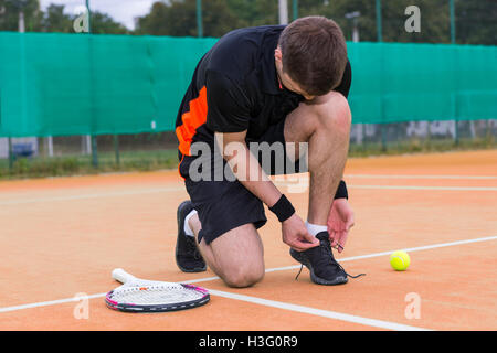 Handsome male tennis player tying shoelaces wearing a sportswear left racket and ball on a clay court outdoor in summer or sprin Stock Photo