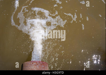 Water flowing from rusted metal culvert drain pipe opening and splashing into foamy brown water, viewed from above. Stock Photo