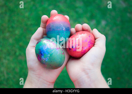 A child's hands holding three dyed, vibrant eggs with metallic accents with a blurred green grass background. Stock Photo