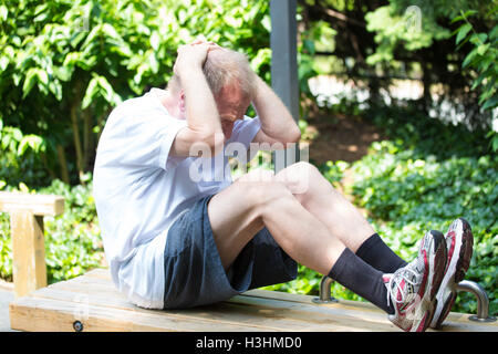 Closeup portrait, healthy elderly man doing sit-ups on bench outside, isolated green trees background Stock Photo
