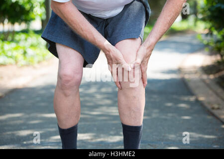 Closeup cropped portrait, older man in white shirt, gray shorts, standing on paved road, in severe knee pain, isolated trees Stock Photo