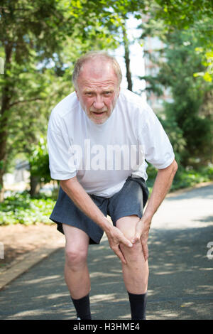 Closeup portrait, older man in white shirt, gray shorts, standing on paved road, in severe knee pain, isolated trees Stock Photo