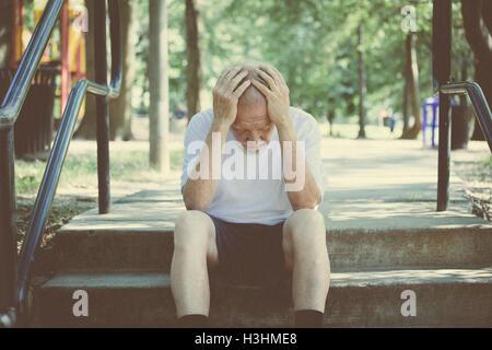 Closeup portrait, senior mature man in black shorts and white shirt, sitting down outside on steps, hands on head in defeat Stock Photo