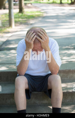Closeup portrait, senior mature man in black shorts and white shirt, sitting down outside on steps, hands on head in defeat Stock Photo